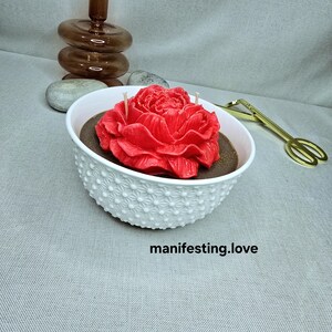 Könnte beinhalten: Eine rote, rosenförmige Kerze in einer weißen, strukturierten Schale. Die Schale enthält eine dunkle Substanz und drei Dochte. Der Text "manifesting.love" ist darunter gedruckt.