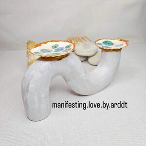 May include: A white ceramic sculpture with a curved, abstract design. Two small, shell-shaped dishes with blue and green painted details sit atop the sculpture. The text "manifesting.love.by.arddt" is visible on the base.