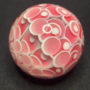 May include: A decorative glass sphere with a pattern of pink and white circular shapes. The sphere has a glossy finish and is set against a dark background. The design features overlapping circles in varying sizes.