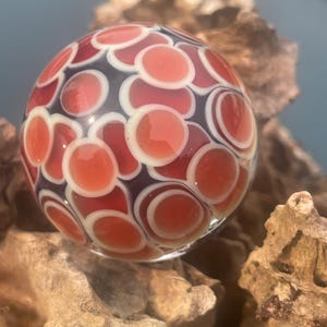 May include: A spherical glass marble featuring a repeating pattern of translucent red circular shapes outlined in opaque white, set against a dark, deep purple background. The surface is smooth and reflective, showcasing a complex, layered design.