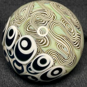 May include: A glass orb with a swirling pattern in white and tan over a pale green base. The orb has several black and white eye-shaped designs on its surface, creating a unique visual texture.