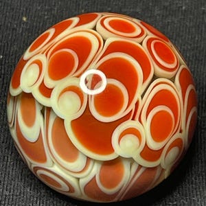 May include: A handcrafted glass marble with a vibrant design. The marble features a pattern of orange and cream-colored swirls and circles, creating a unique and eye-catching appearance. The glass is smooth and rounded.