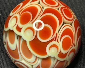May include: A handcrafted glass marble with a vibrant design. The marble features a pattern of orange and cream-colored swirls and circles, creating a unique and eye-catching appearance. The glass is smooth and rounded.
