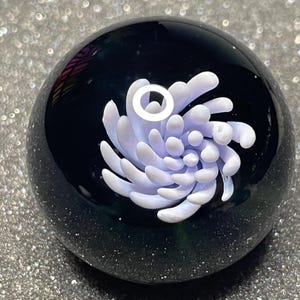 Peut inclure: Un presse-papier sphérique en verre avec une base sombre et mouchetée et un motif floral central violet clair et blanc. Le verre est clair, permettant de voir le motif sous tous les angles. L'objet est décoratif.