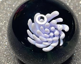 May include: A spherical glass paperweight with a dark, speckled base and a central design of a light purple and white floral pattern. The glass is clear, allowing the design to be visible from all angles. The paperweight is a decorative item.