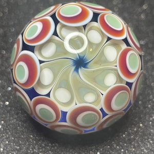 May include: A spherical glass marble featuring a central swirling pattern of pale yellow and blue petals surrounded by an outer layer of repeating circular motifs in shades of orange, white, and light green against a deep blue background.