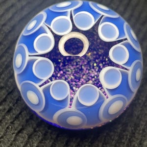 May include: A spherical glass marble featuring a deep blue base with a central star-shaped pattern filled with purple glitter. The surface is decorated with a repeating white and light blue circular motif that radiates from the center point.