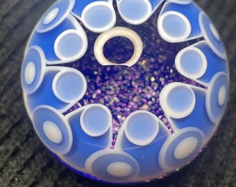 May include: A spherical glass marble featuring a deep blue base with a central star-shaped pattern filled with purple glitter. The surface is decorated with a repeating white and light blue circular motif that radiates from the centre point.