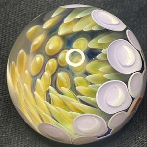 May include: A clear glass paperweight with an abstract design. The interior features yellow, green, and lavender colors arranged in a swirling pattern. A white ring is visible in the center of the paperweight.