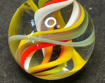 Pode incluir: Uma bolinha de gude de vidro transparente com fitas giratórias amarelas, vermelhas, brancas e verdes. As faixas coloridas criam um padrão em espiral dentro da esfera transparente. Um pequeno círculo branco opaco é visível.