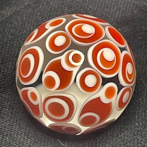 May include: A round, glass marble with a pattern of red and white concentric circles on a gray background. The marble has a clear, glossy finish, and the design is evenly distributed across its surface.