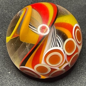May include: A clear glass marble with a vibrant design. The marble features swirling patterns of red, yellow, orange, and black, with white and brown circular accents. The design is intricate and eye-catching.