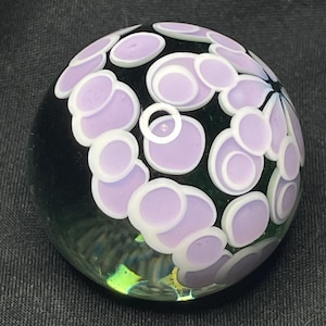 May include: A round glass paperweight with a black base and a pattern of lavender and white circles. The circles are arranged in a floral design, creating a decorative and artistic piece. The paperweight is a unique and eye-catching item.