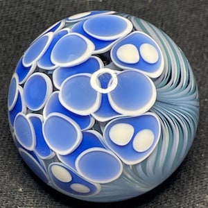 May include: A round glass paperweight with a blue and white floral design. The paperweight features a pattern of blue, oval-shaped elements with white accents, and a swirling blue and white pattern on one side.
