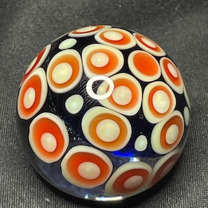 May include: A clear glass paperweight with a dark blue base. The paperweight is decorated with a pattern of orange and white concentric circles. The circles are evenly spaced across the surface, creating a visually appealing design.