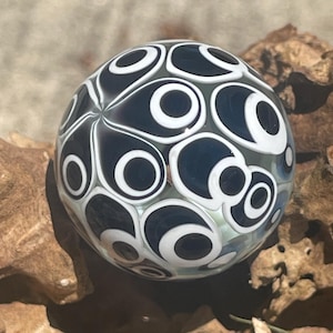 May include: A spherical glass marble featuring a repeating pattern of dark navy blue teardrop shapes outlined in thick white rings. The surface is smooth and glossy, with the intricate design layered within the clear glass orb, creating depth.