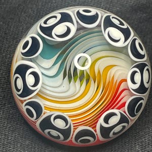 May include: A round glass marble with a swirling rainbow design. The center features a white spiral, surrounded by layers of blue, green, yellow, orange, and red. The marble is encircled by black and white abstract shapes.