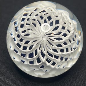May include: A clear glass paperweight with a white, intricate, floral-like design. The design is raised and creates a textured surface. The paperweight has a circular shape and is set against a dark background.