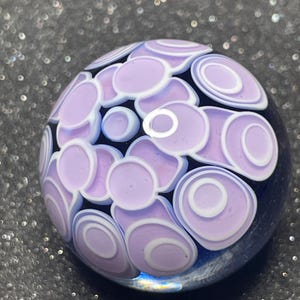 May include: A spherical glass marble with a dark blue base, adorned with a pattern of lavender and white concentric circles and ovals. The design creates a floral or abstract effect. The marble is set against a dark, glittery background.