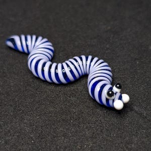 May include: A glass worm figurine with a blue and white striped body. The worm has black eyes and white bulbous features. The figurine is on a dark gray surface.
