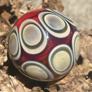May include: A spherical glass paperweight featuring a deep red base color with a series of layered, circular patterns in shades of cream, light blue, and iridescent gold. The surface is smooth and reflective, showcasing a complex, multi-layered design.