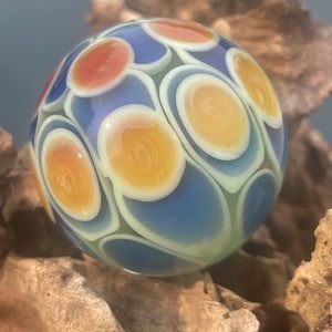 May include: A spherical glass marble featuring a repeating pattern of circular motifs in shades of orange, yellow, and white, set against a translucent blue background. The surface is smooth and reflective, showcasing layered glass work.