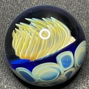 May include: A round glass paperweight with a dark blue background. The paperweight features a yellow and white floral design in the center, with blue and white bubble-like shapes along the bottom edge. The glass is clear, allowing light to pass through.