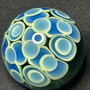 May include: A glass marble with a dark green base and a pattern of blue and light green circles. The circles are arranged in a layered design, with a small white circle at the center of the marble.