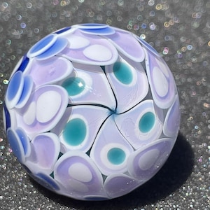 May include: A spherical glass paperweight featuring a layered floral pattern. The design consists of overlapping teardrop-shaped petals in shades of light purple, white, and deep blue, with central accents of teal green arranged in a swirling vortex.