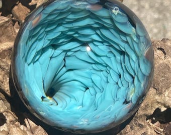 May include: A spherical glass paperweight featuring an intricate internal pattern of layered, light blue, petal-like shapes that spiral toward a dark center. The clear glass exterior creates a magnifying effect over the dense, swirling blue design.