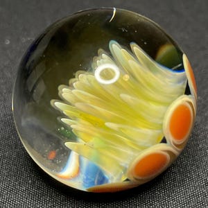 May include: A clear glass paperweight with a yellow, orange, and blue floral design. The interior features a cluster of yellow petals with orange accents and a blue base. The paperweight is round and sits on a dark surface.