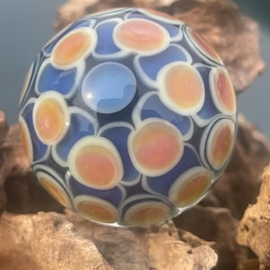 May include: A spherical glass marble featuring a repeating pattern of translucent blue circular shapes surrounded by white rings and centered with iridescent peach-colored discs. The surface is smooth and reflective, showcasing a complex layered design.