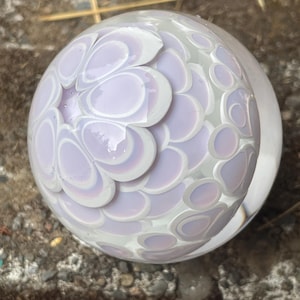 May include: A clear glass paperweight with a spherical shape. The interior features a pattern of overlapping, petal-like shapes in shades of lavender and white. The design creates a layered, three-dimensional effect, with a glossy finish.