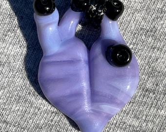 Puede incluir: Un colgante de corazón de vidrio morado con cuentas de vidrio negras en un cordón negro. El corazón tiene un patrón de espiral.