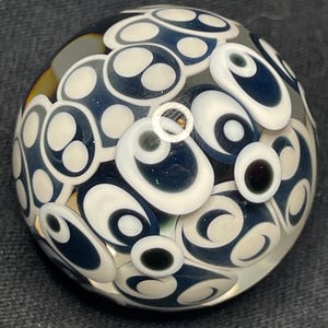 May include: A clear glass paperweight with a pattern of white and navy blue oval shapes. The shapes are arranged in a repeating pattern across the surface of the paperweight. Some shapes have a black center.