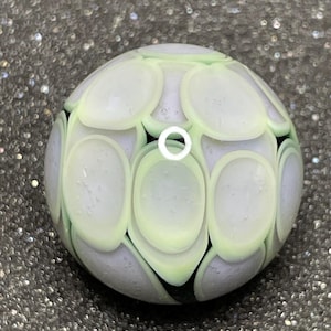 May include: A round, glass bead with a light green and white pattern. The bead has a series of oval shapes in light green and white, set against a lavender background. The bead is a decorative item.