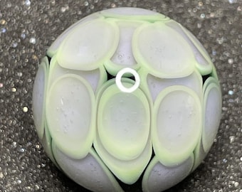May include: A round, glass bead with a light green and white pattern. The bead has a series of oval shapes in light green and white, set against a lavender background. The bead is a decorative item.