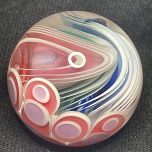 May include: A clear glass paperweight with a swirling design of red, white, blue, and green colors. The paperweight has a series of pink and white circles on the bottom. The design is abstract and artistic.