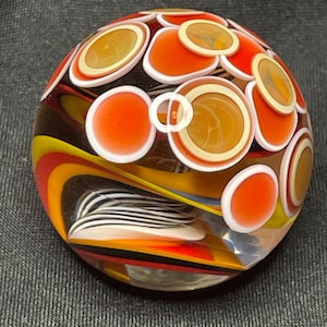 May include: A colorful glass paperweight with a round shape. The paperweight features orange, yellow, and white circular designs on the surface. Swirls of color are visible within the clear glass.