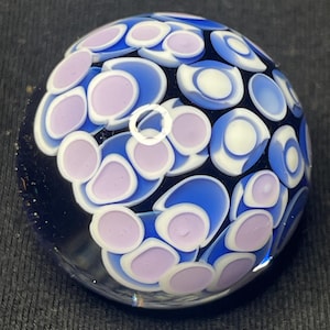 May include: A clear glass paperweight with a dark blue base. The surface is decorated with a pattern of blue, white, and lavender-colored circles. The circles are arranged in a repeating pattern across the surface of the paperweight.
