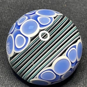 May include: A round glass paperweight with a blue and white design. The paperweight features blue oval shapes with white outlines and diagonal stripes in black, gray, and teal. A small white circle is in the center.