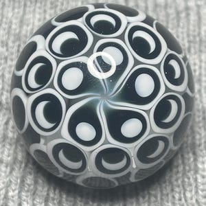 May include: A round, glass marble with a dark teal and white pattern. The design features a series of white, crescent-shaped elements set against a dark teal background. The marble has a glossy finish.