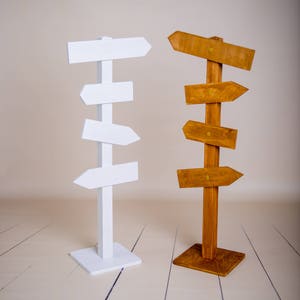 May include: Two wooden signposts with blank arrows. The signpost on the left is white and has four arrows. The signpost on the right is brown and has five arrows.