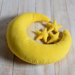 Puede incluir: Una almohada amarilla en forma de media luna con tres almohadas amarillas en forma de estrella encima. Las almohadas están sobre una superficie de madera clara.