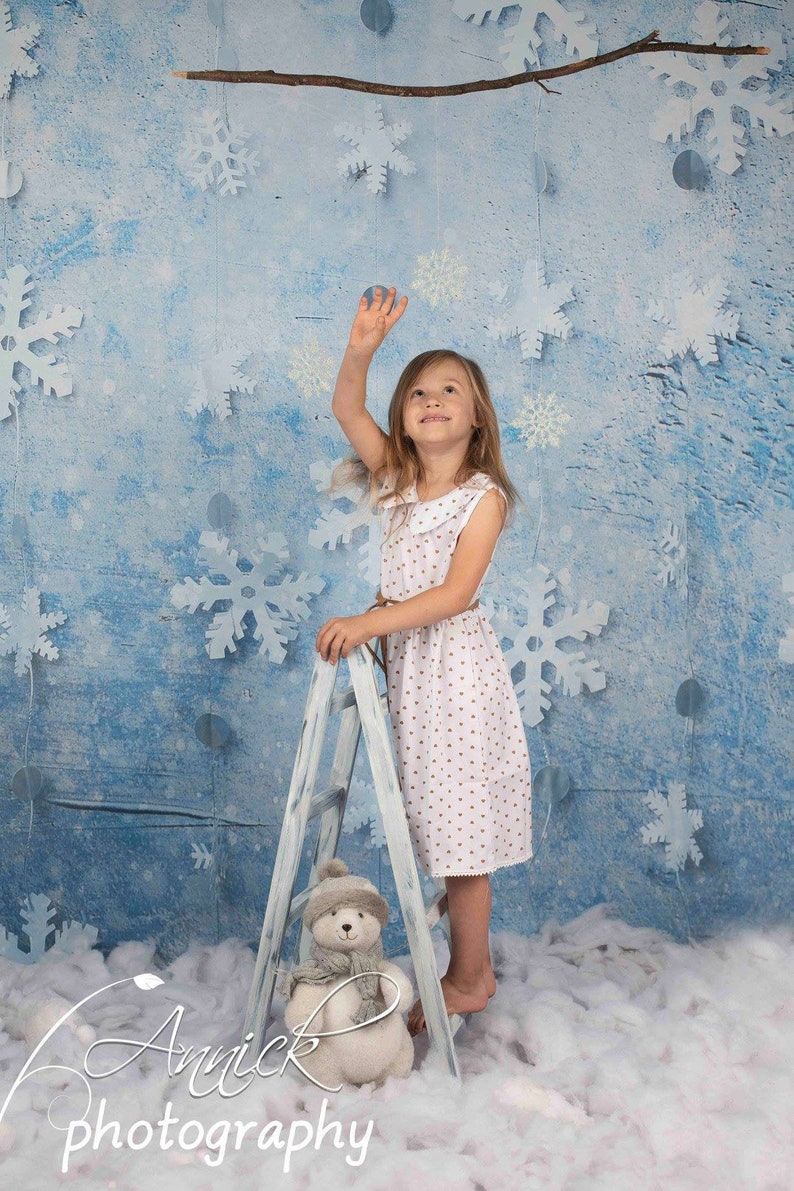 May include: A young girl in a white dress with brown polka dots stands on a white wooden ladder. She is reaching up towards a branch with white snowflakes hanging from it. A white teddy bear with a gray scarf is sitting on the bottom step of the ladder. The background is a blue and white winter scene with snowflakes.
