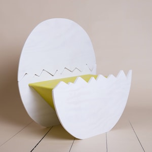 May include: A whimsical, modern chair shaped like a cracked eggshell. The chair is primarily white with a bright yellow seat. The edges of the eggshell are cut in a zig-zag pattern. The chair is made of wood.