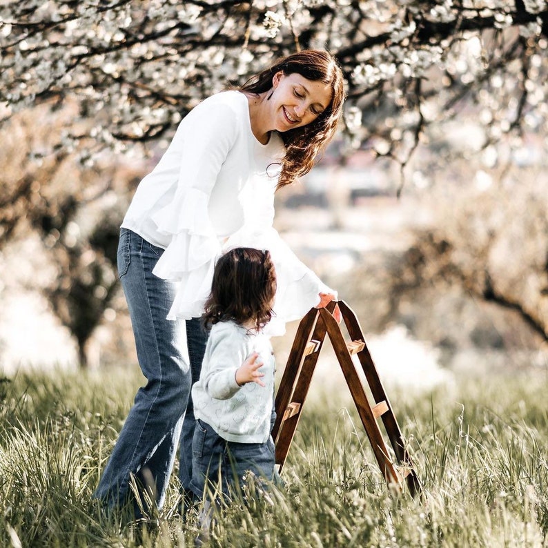 May include: A woman and a child stand in a grassy field with a wooden ladder. The woman is wearing a white long-sleeved shirt and blue jeans. The child is wearing a white long-sleeved shirt and blue jeans. The ladder is made of two pieces of wood that are connected at the top. The ladder is leaning against a tree.
