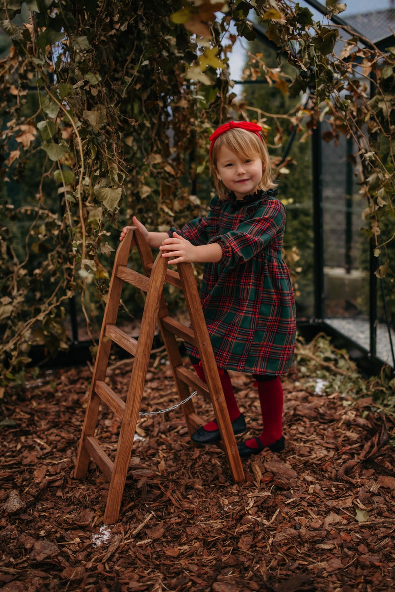 May include: A young girl wearing a red headband and a green and red plaid dress stands next to a wooden step ladder. The ladder has a chain attached to it. The girl is standing on a bed of brown wood chips.