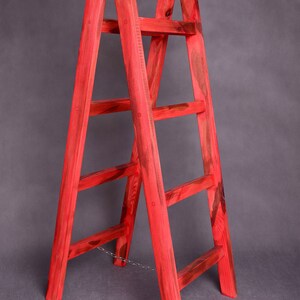 May include: A red wooden ladder with five rungs and a chain connecting the two sides. The ladder is standing on a gray surface.