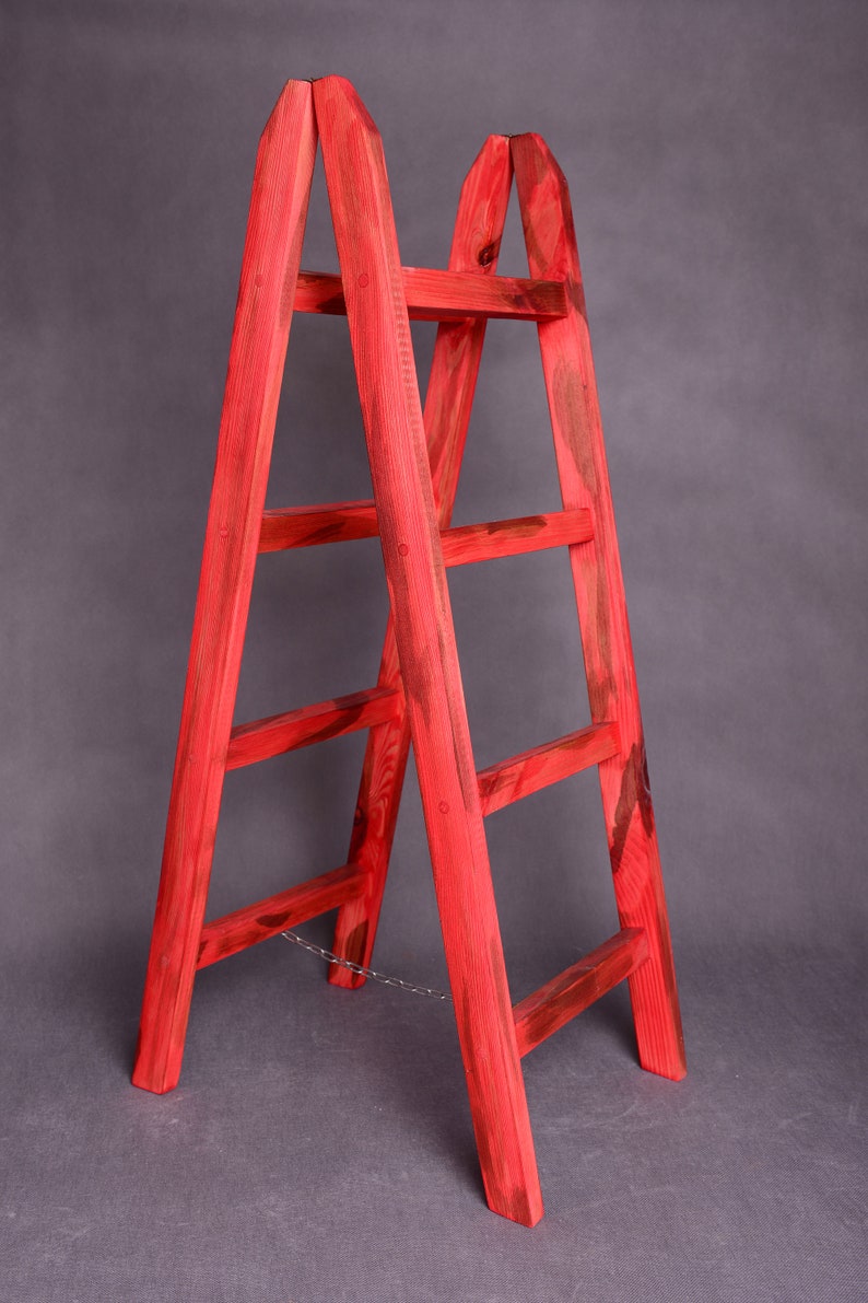 May include: A red wooden ladder with five rungs and a chain connecting the two sides. The ladder is standing on a gray surface.